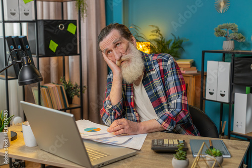 Tired exhausted senior old man freelancer analyzing paperwork graphs, accounting payments and calculating monthly expenses bank bills at home office desk. Upset grandpa working remote job at table.