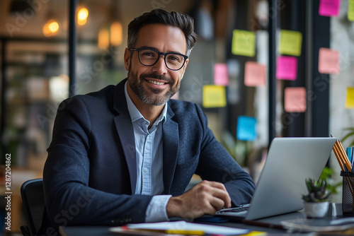 Smiling businessman in a suit working in a modern office with laptop and tablet. Surrounded by colorful sticky notes on a glass wall, brainstorming ideas, planning projects, and boosting productivity.