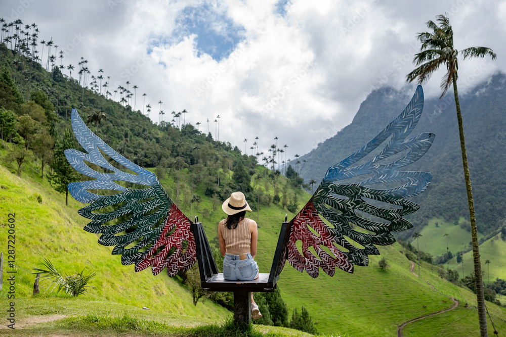 Naklejka premium A woman in casual attire and a straw hat sits on a colorful winged bench overlooking the lush green hills and iconic wax palms of Valle del Cocora in Colombia under a partly cloudy sky.