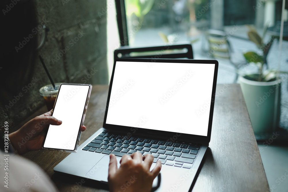 Person using a laptop and smartphone with blank screens, with coffee nearby, in a modern indoor cafe scene. Person using a laptop and smartphone with blank screens, with coffee nearby, in a modern indoor cafe scene.