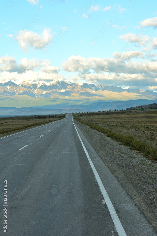 Naklejka premium A two-lane paved road crosses a wide steppe towards a high snow-covered mountain range in the clouds on an early summer morning.