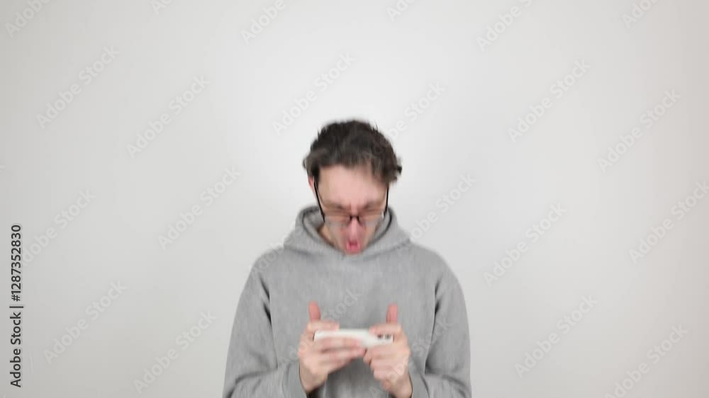 young man, geek in gray hoodie, glasses actively playing and winning computer game on mobile phone by giving thumbs up at the end. isolated on white background. Concept - mobile applications, winner