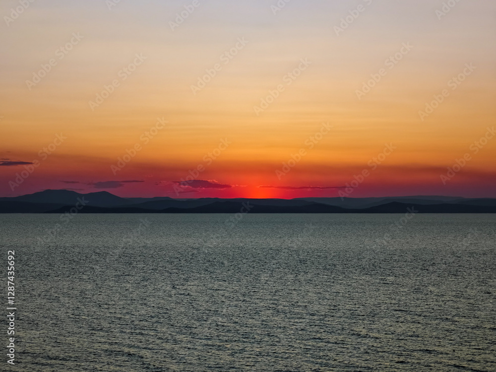Fototapeta premium A breathtaking sunset paints the sky in warm hues over a calm ocean. the horizon is adorned with distant mountain silhouettes, creating a peaceful and serene scene. the blend of colors and natural