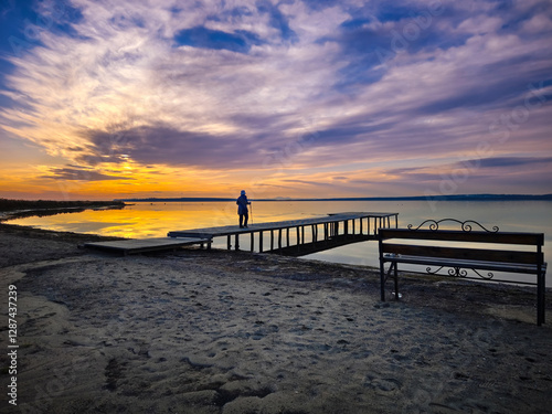 A lone figure stands on a wooden dock, gazing at the vivid sunset over a tranquil lake. the sky's mix of warm oranges and cool purples paint a peaceful scene, enhanced by the sandy beach and a wrought