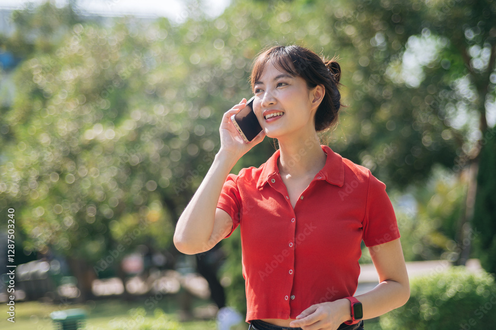 Asian cheerful woman in red shirt talks on her smartphone while standing outdoors in sunny park Asian cheerful woman in red shirt talks on her smartphone while standing outdoors in sunny park