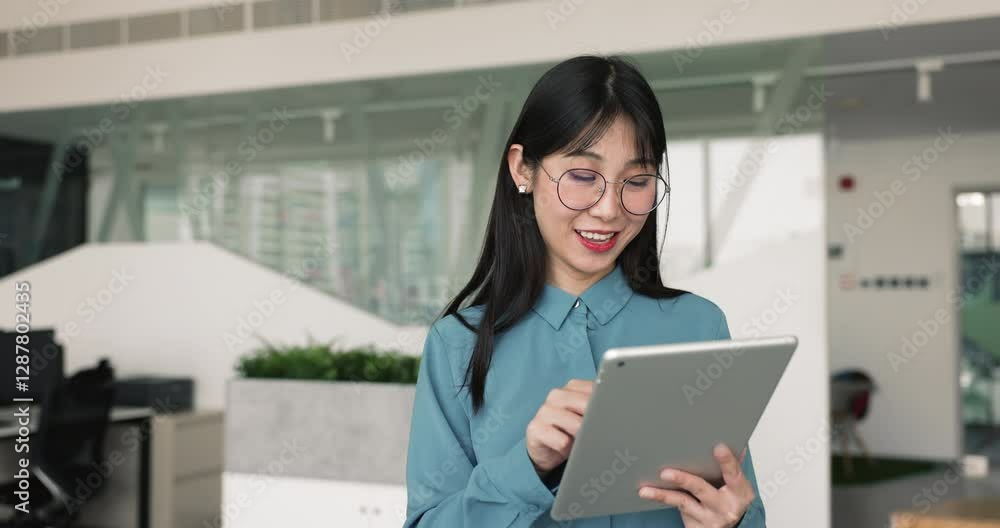 Happy smart young Asian project manager woman in stylish glasses using digital tablet, standing in office space, turning look from gadget away, thinking, dreaming, smiling Happy smart young Asian project manager woman in stylish glasses using digital tablet, standing in office space, turning look from gadget away, thinking, dreaming, smiling