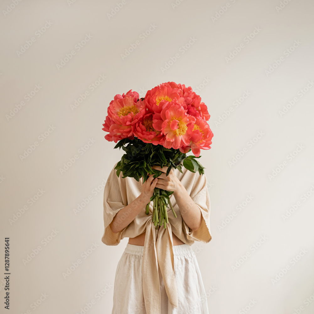 Fototapeta premium Woman in a white dress presents a lush bouquet of vivid red peonies, set against a simple white backdrop. The striking colors and minimalist style. Elegance, romance, and a sense of fresh beauty