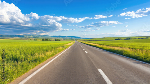 Wide road into distance, flanked by green fields and blue sky. High-definition, horizontal composition. Serene beauty.