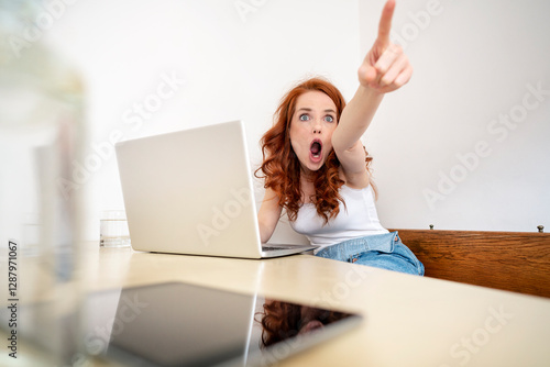 Shocked woman pointing while shouting and sitting with laptop at table against white wall in room