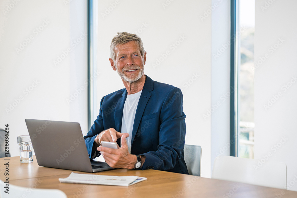 Smiling senior businessman with laptop and cell phone sitting at desk at home Smiling senior businessman with laptop and cell phone sitting at desk at home