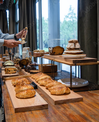 A person selects a pastry from a bakery display case filled with fresh bread and treats. The warm lighting highlights the golden crusts and tempting textures. Perfect for food, bakery, or lifestyle