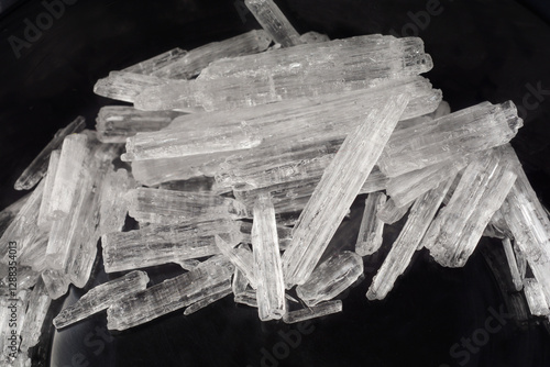 A close-up shot of clear, elongated menthol crystals scattered on a dark surface, highlighting their crystalline texture and reflective properties