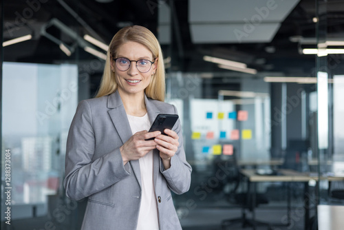 Blonde businesswoman with glasses smiles while holding her smartphone in a modern office setting, ready to tackle the workday