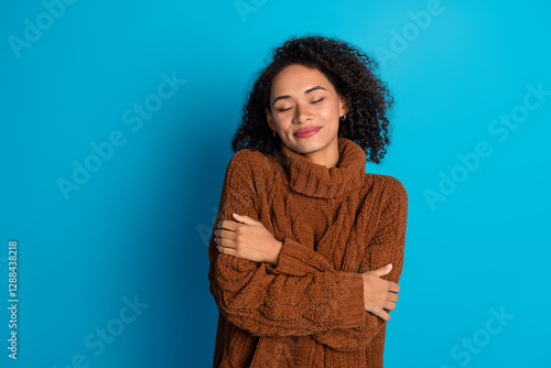 Young woman in a brown sweater embracing herself against a blue background, feeling cozy and content.
