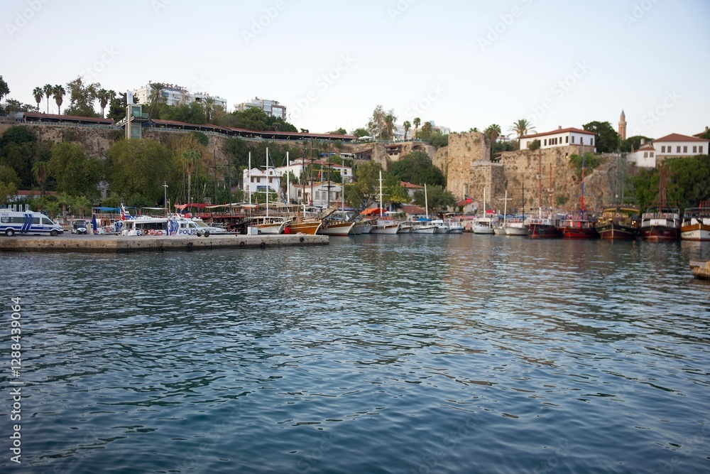 Obraz premium A scenic Mediterranean harbor with various boats docked along the waterfront, historic stone walls, lush greenery, and a coastal town backdrop. Antalya. Turkey 