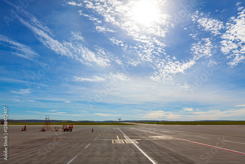 Obraz na płótnie Wide view of an empty airport runway under a bright sky. There are a few pieces of ground equipment and traffic cones visible on tarmac, with vast expanse of open land and distant hills in background w salonie
