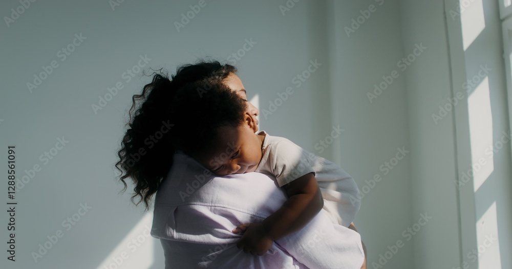 An African-American woman dressed in pastel-colored clothes holds her little daughter in her arms and strokes her back An African-American woman dressed in pastel-colored clothes holds her little daughter in her arms and strokes her back