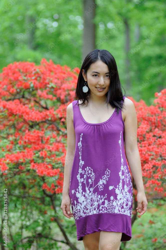 Fototapeta premium Beautiful young Asian woman in purple sundress walks away from bright and blooming shrub in background