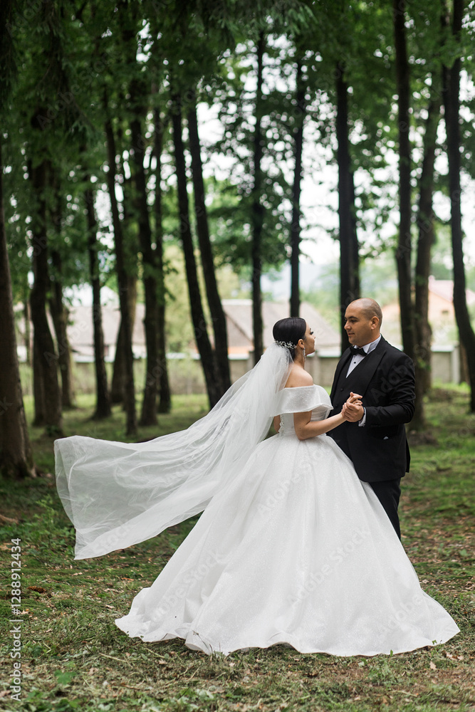 Newlyweds in the forest. The groom is in a black classic suit with a bow tie, and the bride is in a white wedding dress with a long veil. They hold hands and look at each other. Newlyweds in the forest. The groom is in a black classic suit with a bow tie, and the bride is in a white wedding dress with a long veil. They hold hands and look at each other.