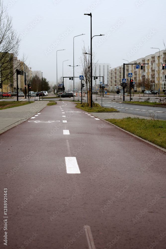 Fototapeta premium Urban street with cycle path and pedestrian walkway surrounded by residential buildings, featuring traffic lights and street signs under a cloudy sky
