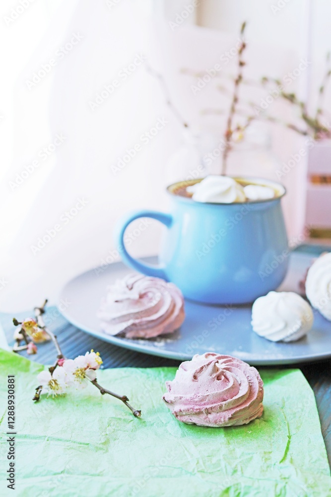 A cup of hot chocolate with marshmallows and a branch of blossoming apricot in a glass vase on a blue wooden table. A cup of hot chocolate with marshmallows and a branch of blossoming apricot in a glass vase on a blue wooden table.