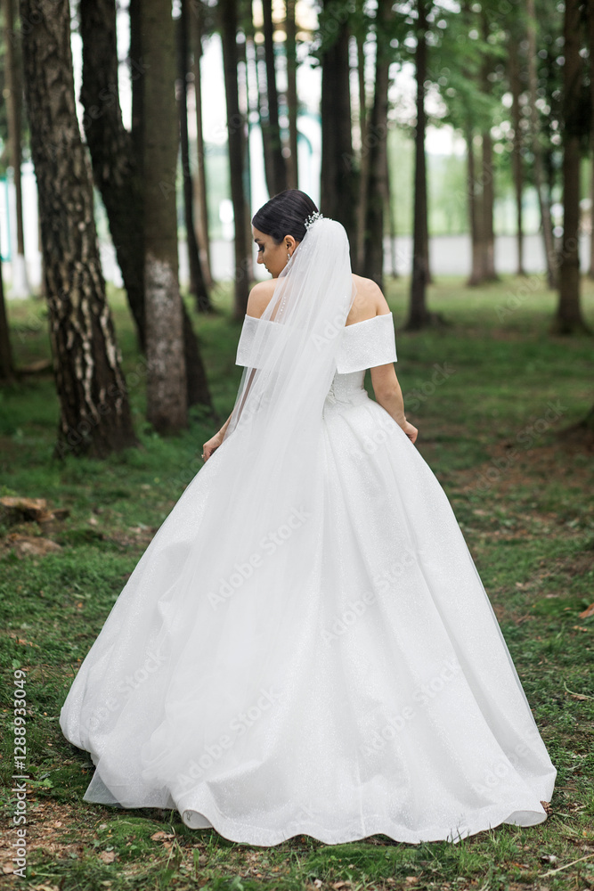A woman in a wedding dress stands in the forest. Her back is turned and her long veil flows down. There are tall trees with dense greenery all around. A woman in a wedding dress stands in the forest. Her back is turned and her long veil flows down. There are tall trees with dense greenery all around.