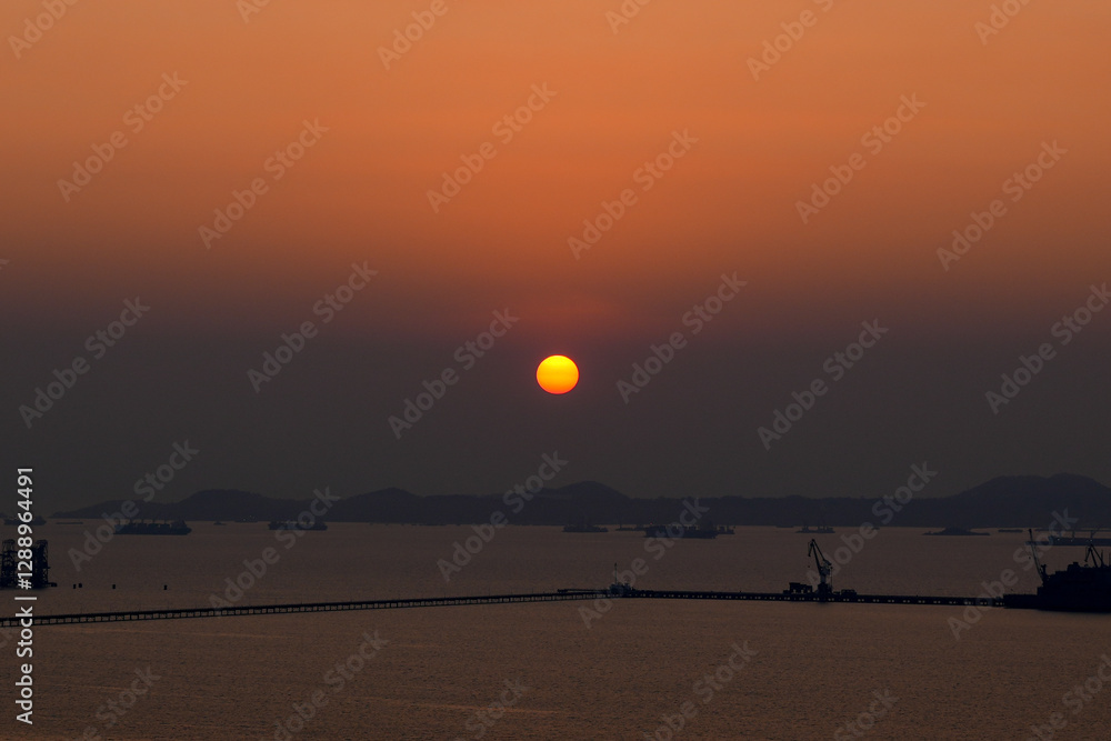 Beautiful sunset over industrial port with silhouettes of cranes and cargo ships in the distance and islands on the horizon Beautiful sunset over industrial port with silhouettes of cranes and cargo ships in the distance and islands on the horizon