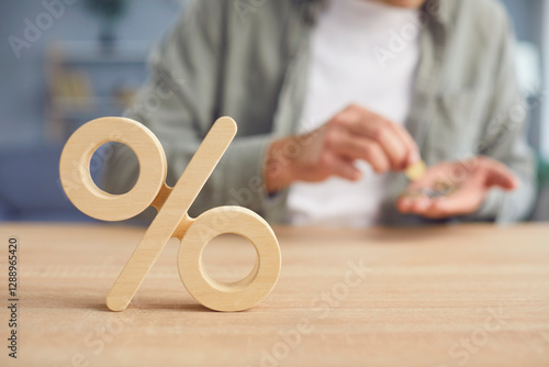 Close up cropped shot of wooden percentage symbol on desk. Young man counting money in his hands in the background. interest rate decline, investment reduce or sales discount concept. Selective focus.