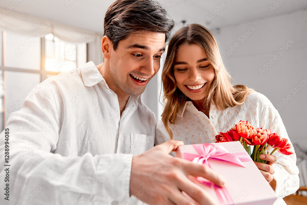 A happy couple in white outfits excitedly opening a pink gift box. The woman holds a bouquet of red tulips, both smiling warmly in a bright, cozy home setting.Joyful Surprise.Couple Unwrapping a Gift. A happy couple in white outfits excitedly opening a pink gift box. The woman holds a bouquet of red tulips, both smiling warmly in a bright, cozy home setting.Joyful Surprise.Couple Unwrapping a Gift.
