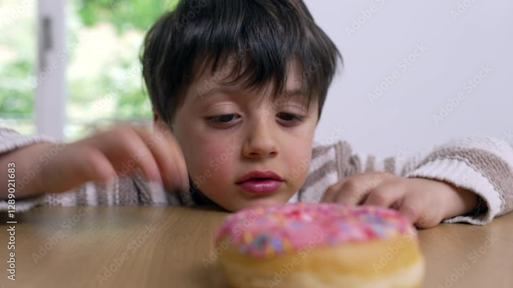 Young boy tempted by a pink frosted donut with colorful sprinkles, anticipation and curiosity in his eyes, staring at it while sitting at a wooden table in a bright, sunlit room