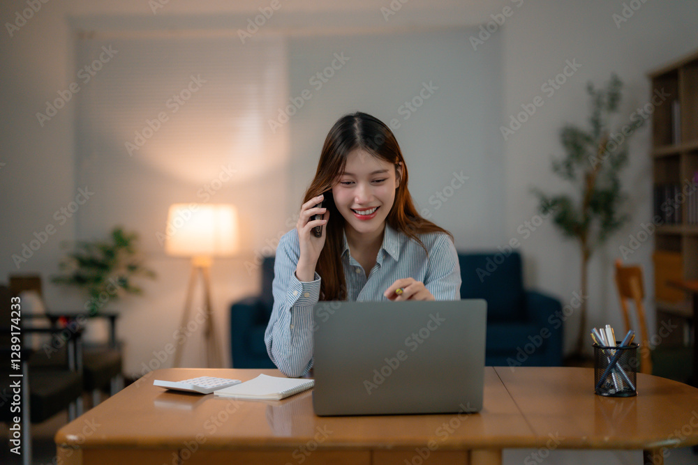 Young professional working remotely, Asian businesswoman smiling while using laptop and mobile phone during late-night home office session, freelancing with confidence Young professional working remotely, Asian businesswoman smiling while using laptop and mobile phone during late-night home office session, freelancing with confidence