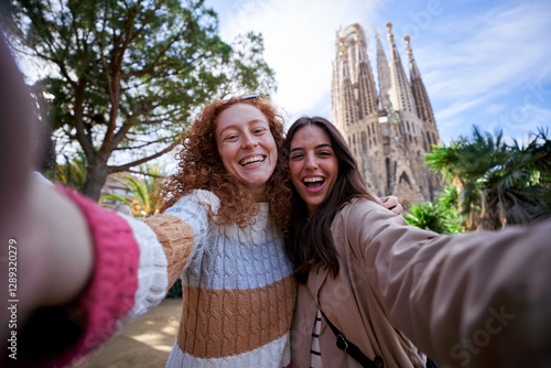 Two happy women friends taking a selfie in front of the sagrada familia, enjoying their vacation together, looking at camera smiling.