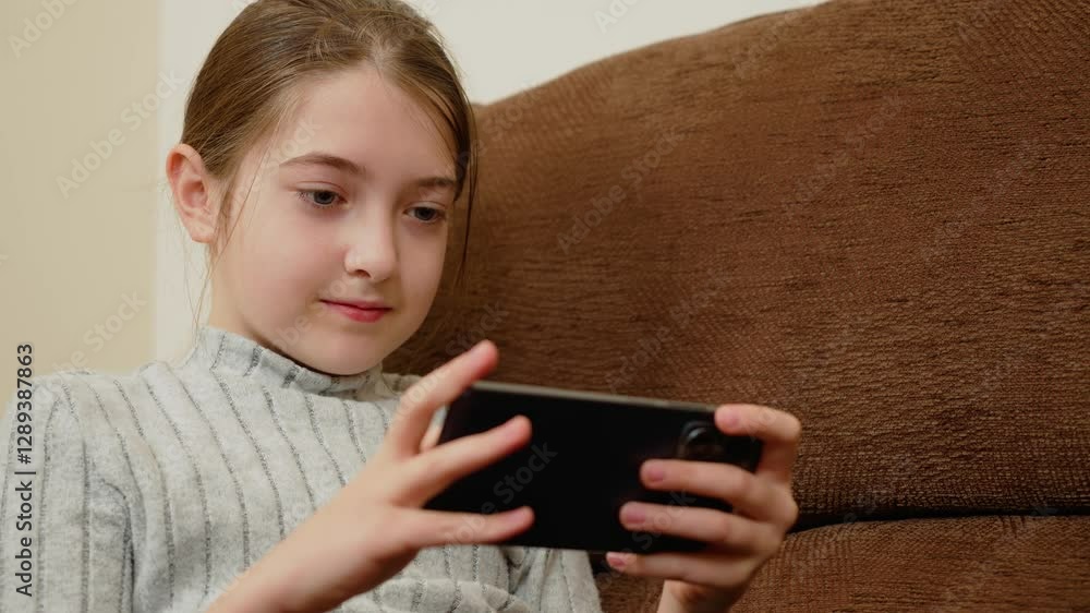 Girl playing on smartphone while sitting on couch. Kid focusing on screen during mobile activity. Child using phone for leisure. Person engaged in digital interaction on device Girl playing on smartphone while sitting on couch. Kid focusing on screen during mobile activity. Child using phone for leisure. Person engaged in digital interaction on device