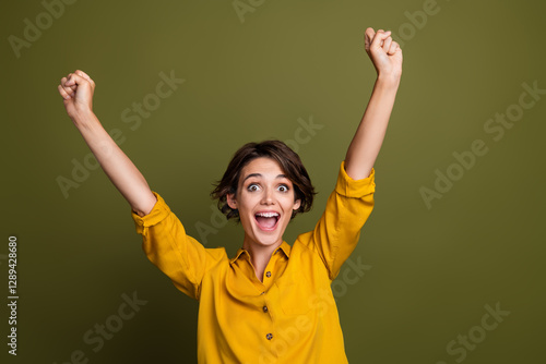 Happy young woman in a yellow shirt celebrating success with arms raised on a khaki background. Styled for casual scenarios. photo