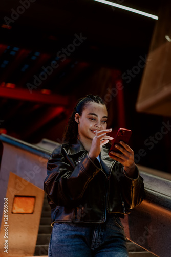 Young Hispanic woman in leather jacket sitting on urban stairs at night, smiling while texting on smartphone. Warm city lights create cozy atmosphere. Enjoying social media and online communication