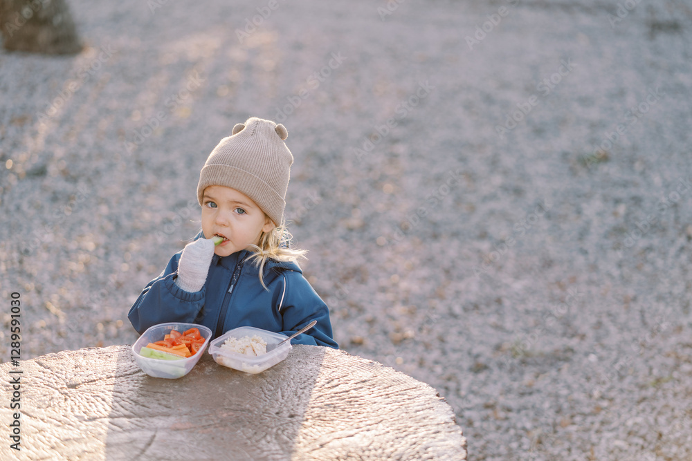 Little girl eats a piece of cucumber while standing behind a large tree stump with a lunchbox Little girl eats a piece of cucumber while standing behind a large tree stump with a lunchbox