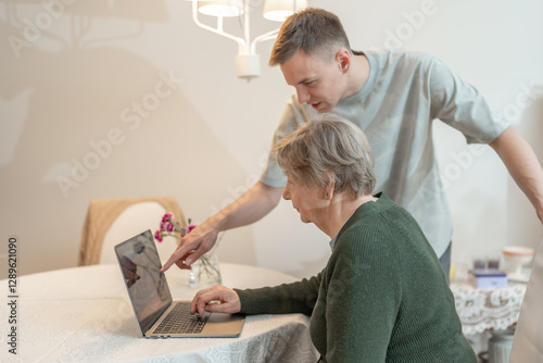 Young man helping an elderly woman with a smartphone at home. Concept of technology, education, and senior digital adaptation. Grandson teaches grandmother how to use digital device.