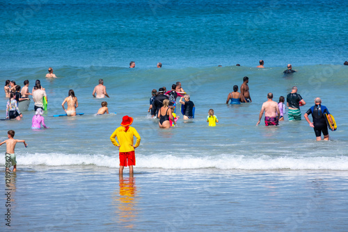 A lifeguard in a bright color uniform stands at the beach, watching numerous people or beachgoers swim and enjoying the waves on a summer day at Great Ocean Road. Torquay, VIC Australia.
