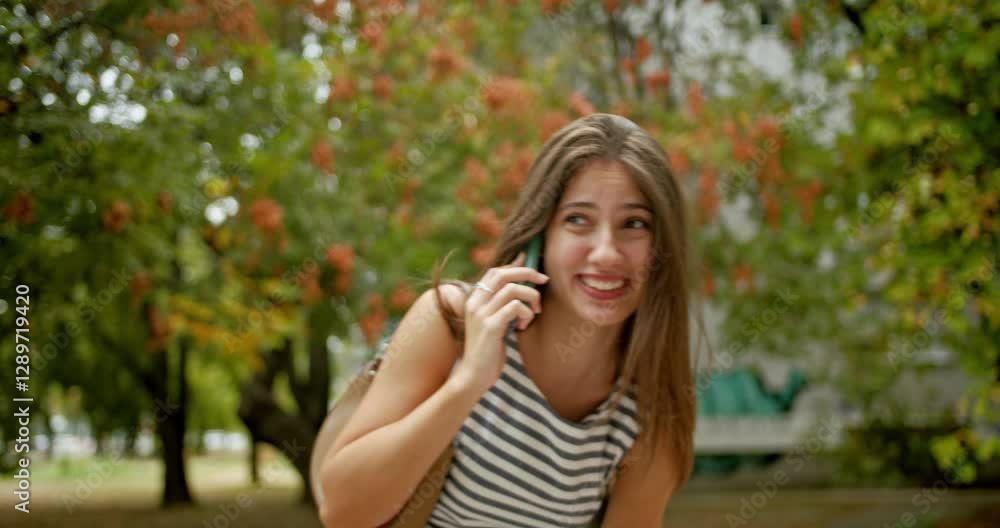 A beautiful young student woman is walking through a park. She's talking on her phone and laughing.
