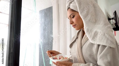 Young Latina woman dressed in a bathrobe and a towel wrapped around her head eats a bowl of fruit with yogurt and oatmeal by the window