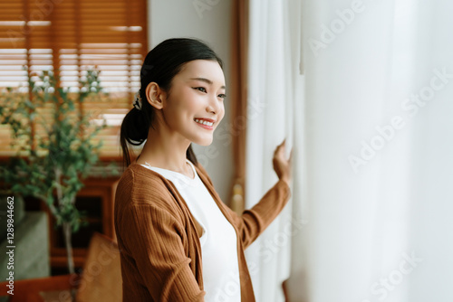 Fotografie Happy Asian woman standing by the window and looking outside the living room enj