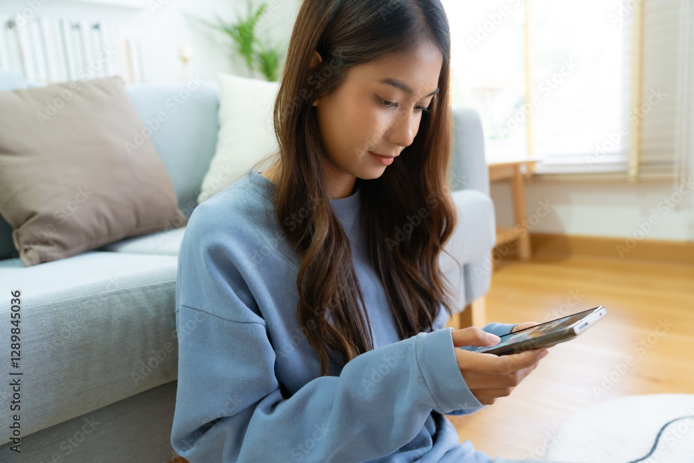 Smiling pretty young Asian woman sitting on cozy couch, using funny mobile apps in living room. Woman at home, Doing online shopping, Messaging friends, Posting on social Media Smiling pretty young Asian woman sitting on cozy couch, using funny mobile apps in living room. Woman at home, Doing online shopping, Messaging friends, Posting on social Media