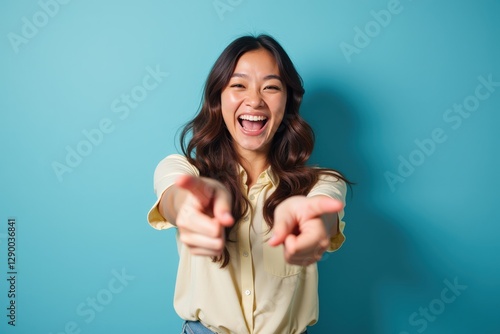 Cheerful Asian Young Woman in Yellow Blouse Smiling and Pointing Playfully Against a Vibrant Teal Background in a Bright, Energetic Setting
