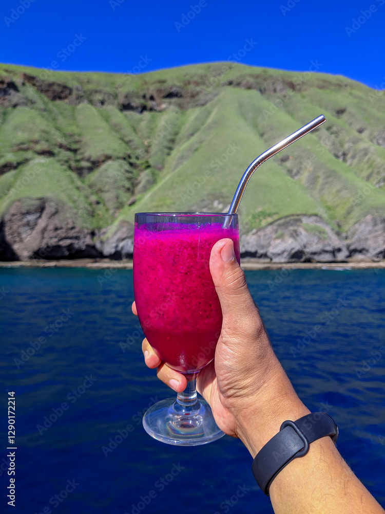 A hand holding a glass of vibrant dragon fruit juice with a metal straw, set against a scenic backdrop of a lush green hill and deep blue ocean under a clear sky