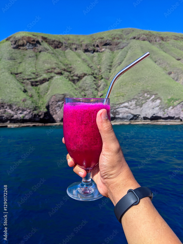 A hand holding a glass of vibrant dragon fruit juice with a metal straw, set against a scenic backdrop of a lush green hill and deep blue ocean under a clear sky