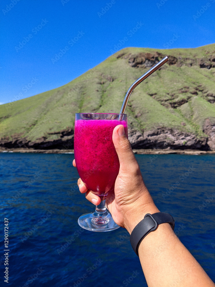 A hand holding a glass of vibrant dragon fruit juice with a metal straw, set against a scenic backdrop of a lush green hill and deep blue ocean under a clear sky