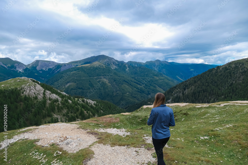 Naklejka premium A woman gazes at the stunning, lush panorama of the Nocky Mountains in Carinthia, Austria. Overlooking the vast, green hills under a cloudy sky, enjoying the scenic view and fresh air