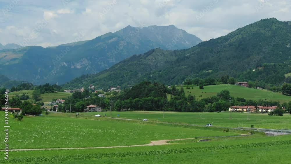Busy road crossing Clusone plateau, green fields and meadows with mountains in background, Rovetta, Italy. Aerial forward ascending Busy road crossing Clusone plateau, green fields and meadows with mountains in background, Rovetta, Italy. Aerial forward ascending