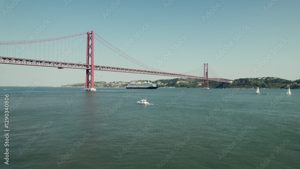 Naklejka premium The ship sails under a red suspension bridge on a wide river in Portugal. Yachts and sailboats sail on the river. Drone video.