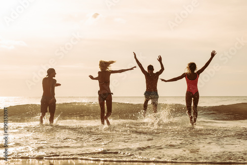 Group of four happy young friends runs to sunset sea beach and jumps over waves with splashes. Tropical vacations concept